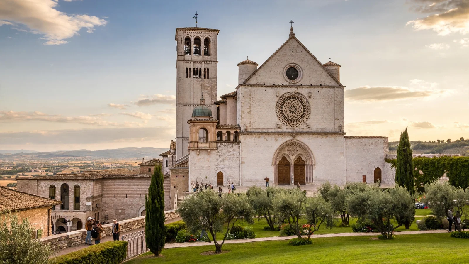 Basilica of St Francis in Assisi, a pale stone Franciscan church with a tall bell tower and a large rose window, seen from a lawn with olive trees and cypresses