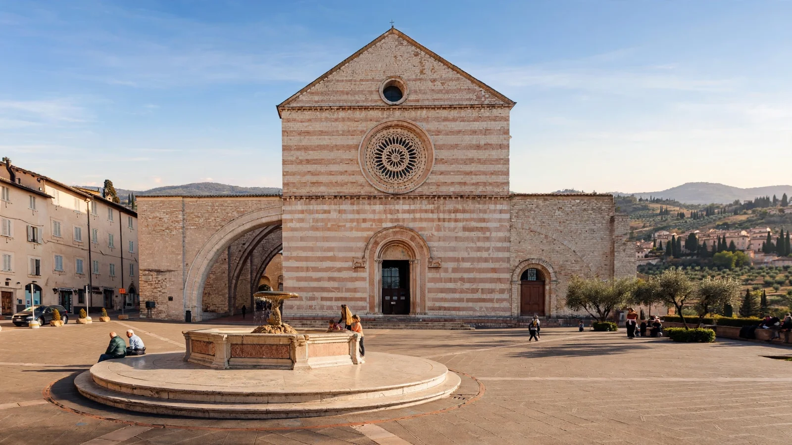 Basilica of Santa Chiara in Assisi, a church with a pink and white striped stone facade, a rose window and large side buttress arches, in a square with a stone fountain