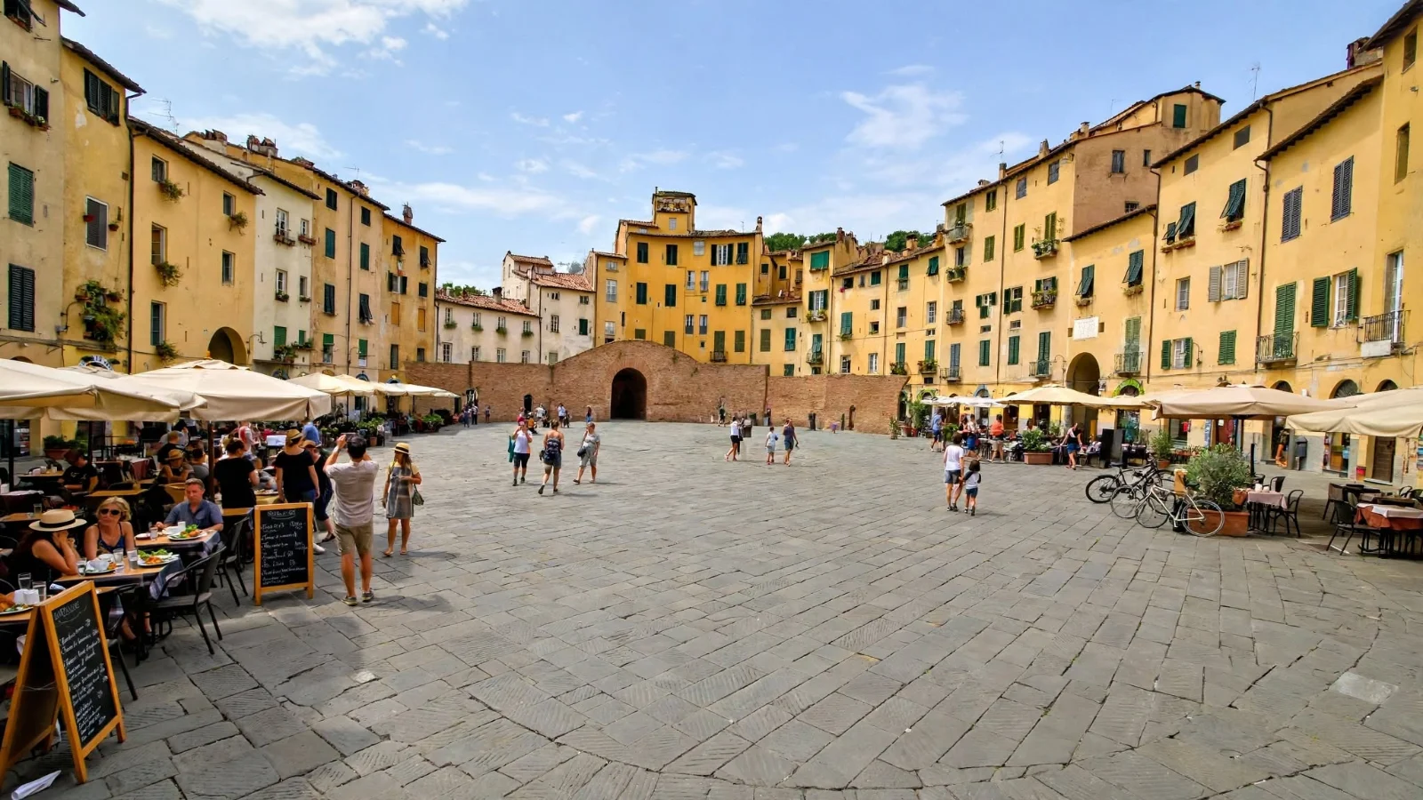 Oval Piazza dell'Anfiteatro in Lucca surrounded by yellow ochre buildings, outdoor cafe umbrellas, a vaulted brick entrance gate, and flat stone slab paving