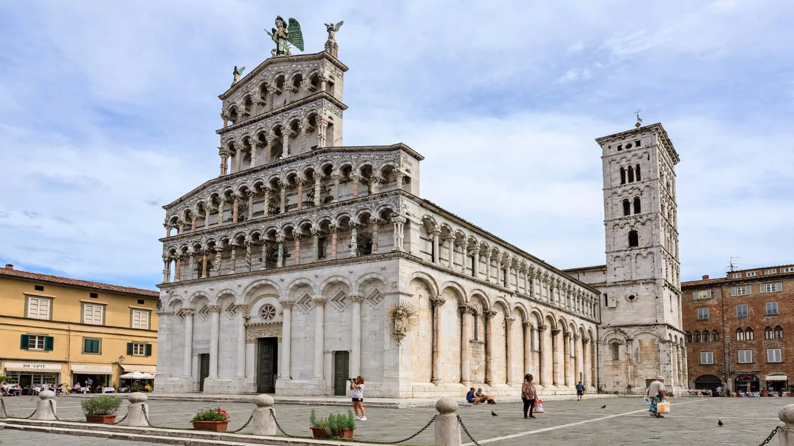Ornate four-tiered white marble facade of San Michele in Foro in Lucca topped by the winged statue of Archangel Michael on stone-paved Piazza San Michele