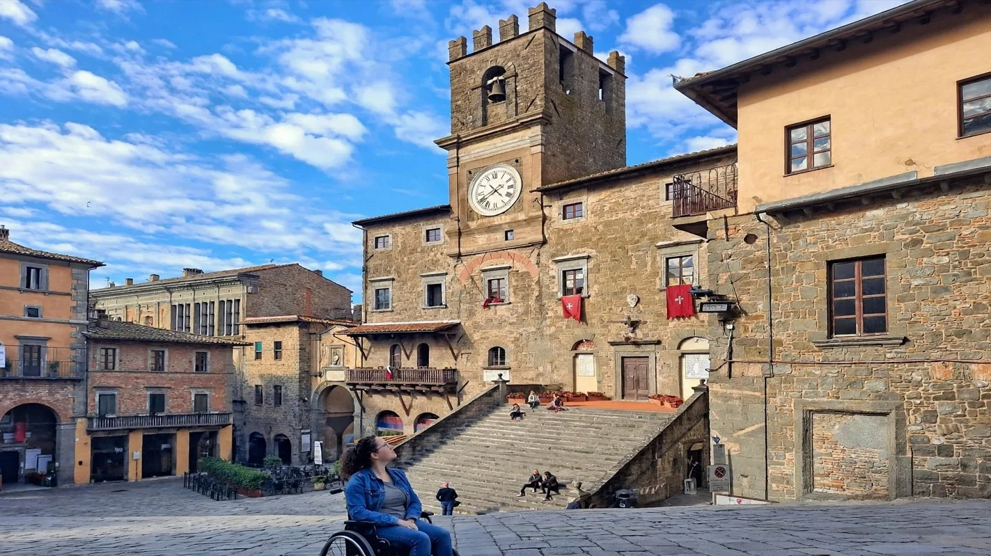 Wheelchair user reaching the courtyard ramp and lift entrance at Palazzo Comunale in Montepulciano, with the narrow tower stairs shown in an inset