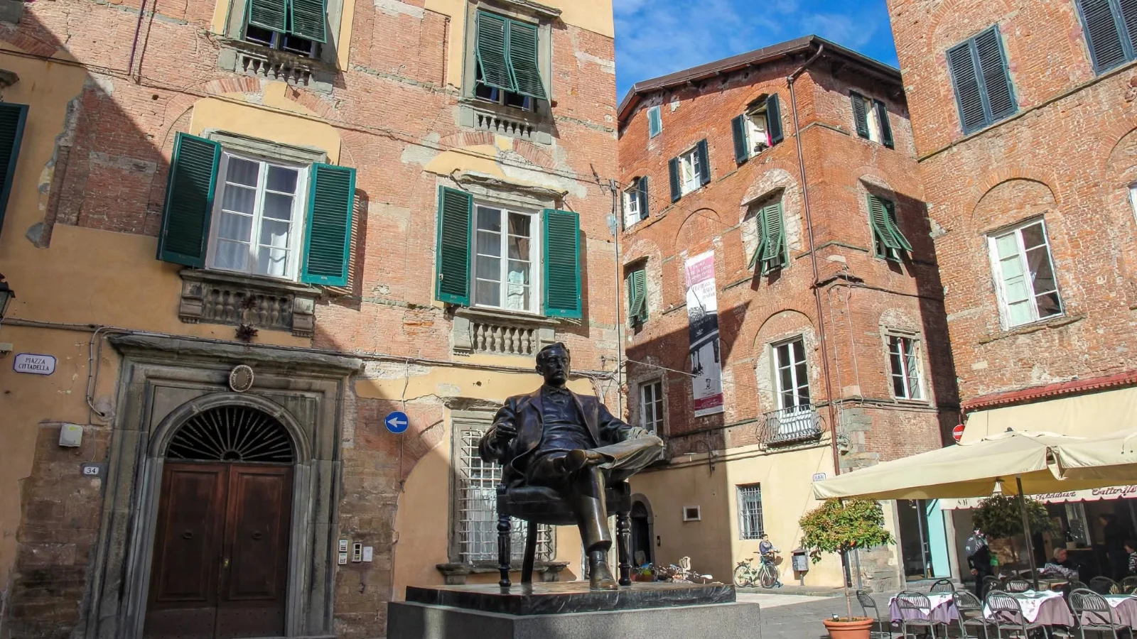 Piazza Cittadella in Lucca showing the seated bronze statue of composer Giacomo Puccini in front of his historic brick birth house with green wooden shutters